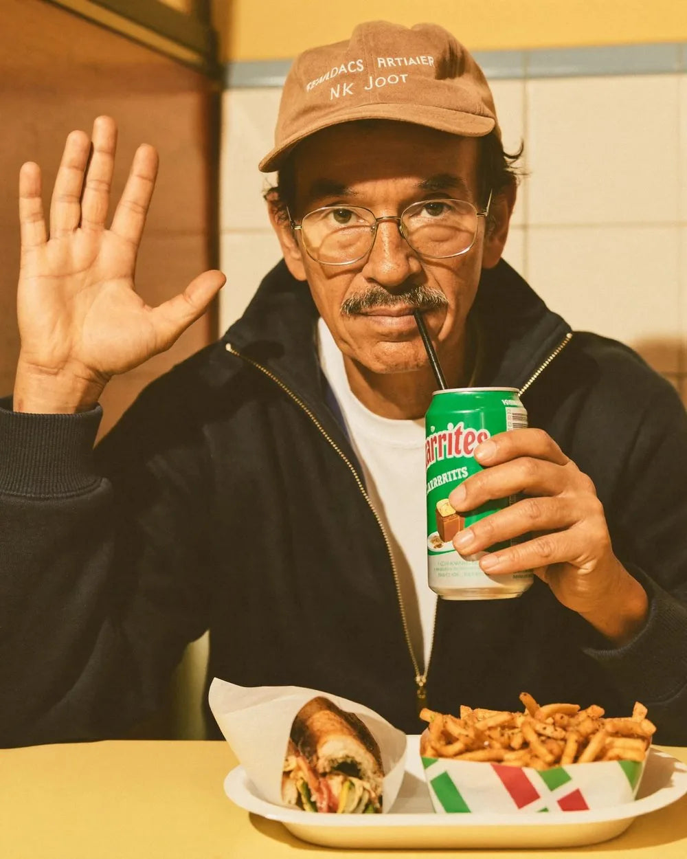 A man in a diner booth waving, with a drink and a tray of food