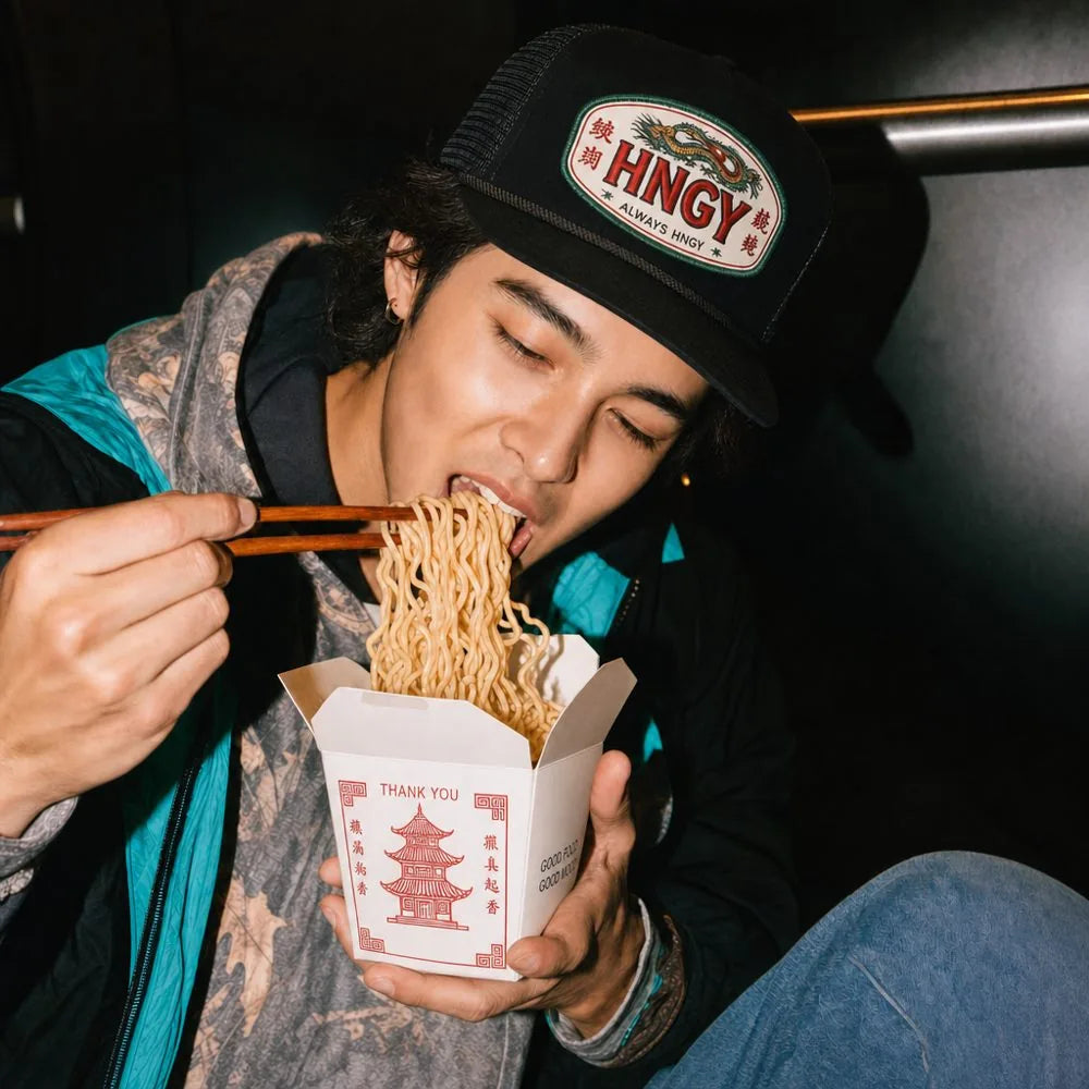 A man in a HNGY cap eating noodles with chopsticks